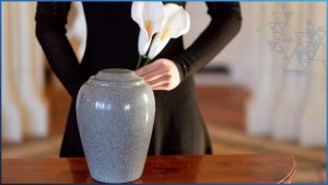 In the foreground is a medium gray funeral cremation remains urn sitting on a medium-tone wooden table. In the background is a torso and arms of a mourning woman wearing black, holding white and yellow flowers.