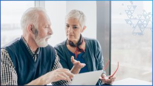 Two senior adults, both wearing muted greenish blue clothes, sit at a table and quietly discuss something. On the left is a man holding a piece of paper. On the right is a woman holding a pair of red reading glasses and gesturing with her other hand. Both people have short white hair.