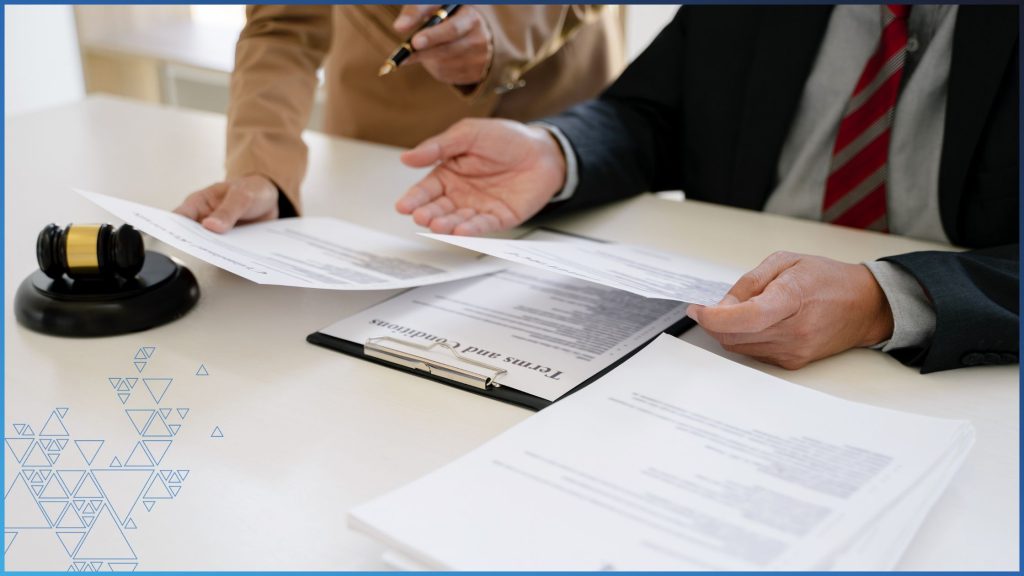 The hands of two businesspeople are shown behind a desk holding legal documents. On left is woman wearing a tan suit. On left is a man wearing a dark navy suitjacket. Each person is holding a sheet of paper with text on it. On a light pine colored desk is a stack of papers on the right and more papers on a clipboard on the left.