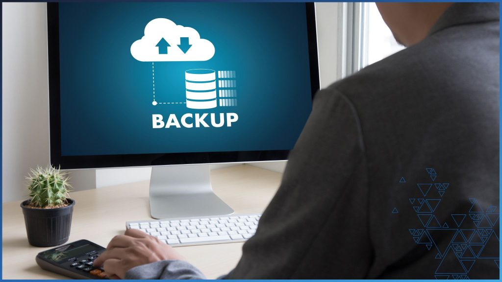 Seen from the back, a man in a dark business suit sits at a desk looking at a computer monitor on which is a teal screen with white graphic indicating cloud backup in progress with the word "Backup." A white keyboard is just in front of the monitor. The man's left hand rests on a calculator. To the left of the computer screen is a small cactus in black pot.