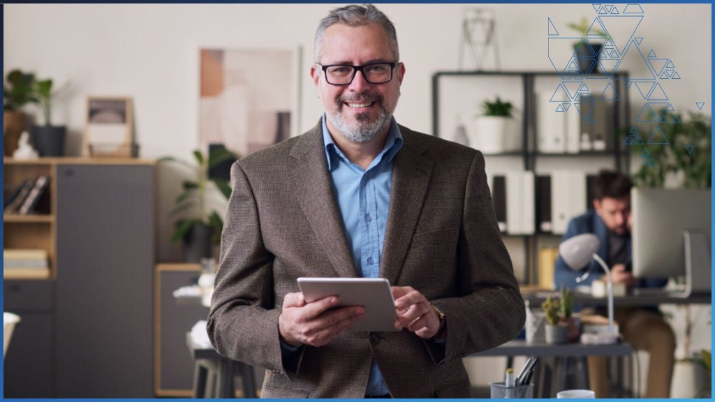 A middle-aged man in a brown suit and blue shirt with glasses stands in an open-plan business office. He is smiling, facing the camera, and holds a computer tablet in his hands. It's implied that he is in the deathcare industry and is using funeral home management software.