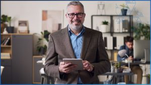 A middle-aged man in a brown suit and blue shirt with glasses stands in an open-plan business office. He is smiling, facing the camera, and holds a computer tablet in his hands. It's implied that he is in the deathcare industry and is using funeral home management software.