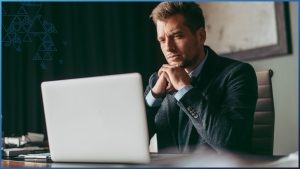 A middle-aged businessman in a gray suit sits at desk, facing the camera, looking at laptop open on his desk, with a serious expression on his face. It's implied that he works in the deathcare industry and is using funeral home software.